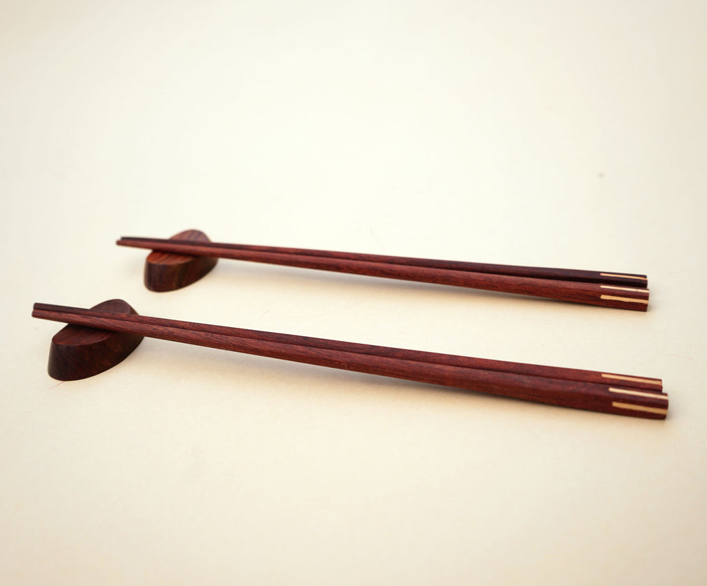 Experience the art of dining with a set of handmade wooden chopsticks with matching rests. Chopsticks and rests are handmade from wood with a natural wooden colour. Finished with a hand polish for a touch of a natural shine. Come in a set of 2 pairs of wooden square chopsticks with 2 wooden rests.   Suitable for hot or cold food. Ideal gift and home use. Handmade and fair trade product. 